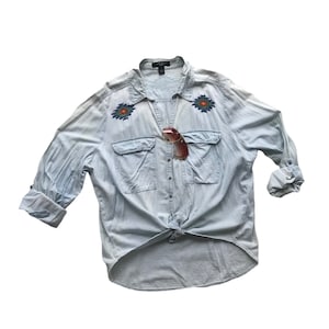 Può includere: Una camicia di jeans blu chiaro a maniche lunghe con un nodo sul davanti. La camicia presenta disegni geometrici ricamati sul petto e una chiusura con bottoni sul davanti.