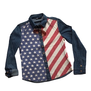 Può includere: Camicia di jeans con un design a bandiera americana. Il lato sinistro presenta uno sfondo blu con stelle bianche, il lato destro ha strisce rosse e bianche. Include occhiali da sole marroni.