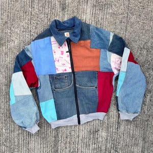 May include: A blue denim jacket with a patchwork design. The jacket has a zipper closure and is made of different shades of blue denim, red, orange, and white fabric.