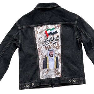Op de afbeelding: Een zwart denim jack met een patch op de rug. De patch heeft een camouflagepatroon, de vlag van de Verenigde Arabische Emiraten en een portret van een man in traditionele kleding.