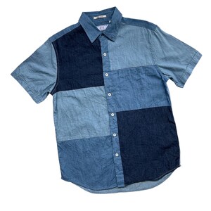 May include: A short-sleeved shirt with a patchwork design in various shades of blue denim. The shirt has a button-down front, a classic collar, and a single chest pocket. The brand name "RVC" is visible on the collar. The shirt is made of denim.