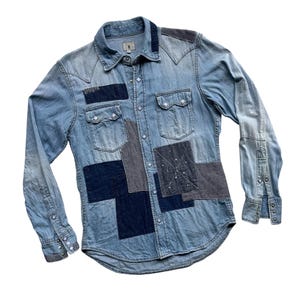 Puede incluir: Camisa vaquera azul claro con detalles de patchwork. La camisa tiene un cuello clásico, cierre de botones en la parte delantera y mangas largas. Parches de tela azul oscuro y a rayas están cosidos en la parte delantera y las mangas. Se ven dos bolsillos en el pecho.