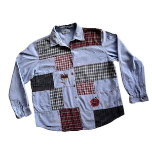 Può includere: Una camicia blu a maniche lunghe in patchwork con un motivo a quadri rosso e nero. La camicia ha una mela ricamata sul davanti.