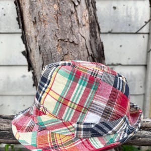 Op de afbeelding: Een patchwork bucket hat met een kleurrijk ruitpatroon. De hoed heeft vierkanten van rode, witte, groene, gele en marineblauwe stof. De hoed ligt op een houten oppervlak.