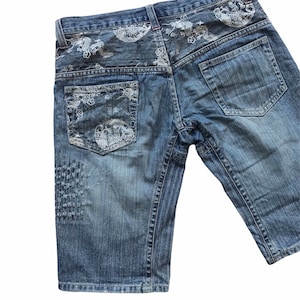 Op de afbeelding: Blauwe denim shorts met een wit geborduurd drakenontwerp op de achterzakken. De shorts hebben een distressed look met verbleekte gebieden en stiksels.