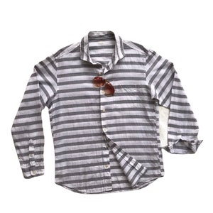 May include: A long-sleeved striped button-down shirt in white and gray with a pocket on the left side. Sunglasses are tucked into the collar.