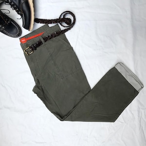 Puede incluir: Un pantalón cargo verde oliva con un cinturón trenzado marrón y un lazo de cinturón rojo. El pantalón tiene un dobladillo blanco en la parte inferior.