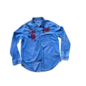 Könnte beinhalten: Ein hellblaues Jeanshemd mit langen Ärmeln und Knopfleiste. Das Hemd ist mit gestickten roten Rosen auf der Vorderseite verziert.