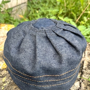 May include: A denim cap with a rounded crown and a brimless design. The dark blue fabric is accented with three rows of tan stitching around the base. The cap has a pleated top.