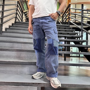 May include: A white short-sleeved t-shirt with a small, colorful patterned chest pocket and blue denim cargo pants featuring various shades of blue fabric patches, frayed edges, and multiple pockets, paired with light gray athletic sneakers.