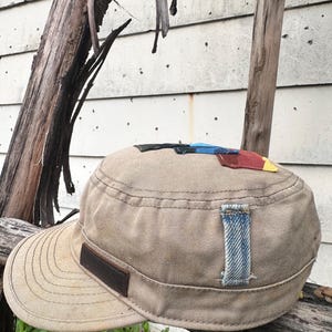 Puede incluir: Gorra de lona color beige con una visera corta y un parche de cuero marrón. La gorra presenta parches de tela coloridos en la parte superior y un parche de mezclilla en el lateral. El sombrero está sobre una superficie de madera.