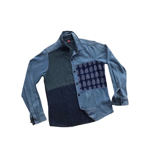 以下が含まれることがあります： パッチワークのデザインが施された青い長袖デニムシャツ。シャツは襟、ボタン留め、ポケットが2つ付いています。パッチワークには、さまざまな色の青いデニムと模様のある生地が使われています。