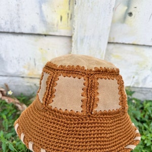 May include: A brown crocheted bucket hat with a suede brim and crown. The hat has a unique design with suede squares sewn into the crown.