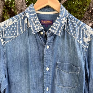 May include: A blue denim button-down shirt featuring a white patterned yoke and collar with floral and geometric designs. The shirt includes a front chest pocket, white buttons, and a purple interior label reading Purple & Yellow with size L.