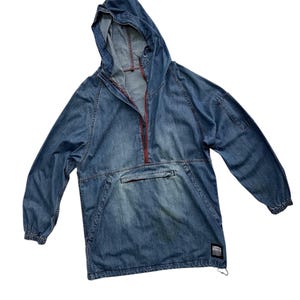 May include: A blue denim hooded anorak jacket with a red zipper and a front pocket. The jacket has long sleeves and a drawstring at the bottom. A small label is visible on the bottom right.