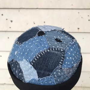 May include: A blue denim patchwork beanie with a black band. The beanie is made of different shades of blue denim and is stitched together with white thread.