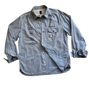 Può includere: Una camicia abbottonata a maniche lunghe di colore azzurro chiaro con una varietà di timbri e disegni stampati in nero. La camicia ha una singola tasca sul petto con patta.