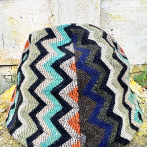 May include: A colourful, patterned hat with a zig-zag design in shades of green, blue, black, orange, and white. The hat is made of a soft, textured fabric.