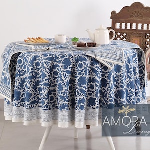 May include: A white table with a blue and white floral tablecloth. The table is set with two cups, a plate of cookies, and a teapot. A wooden chair with a carved back is next to the table.