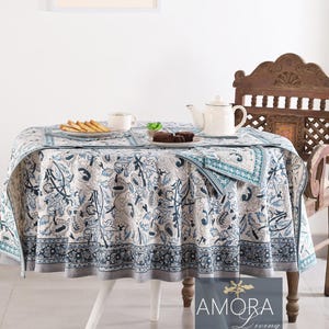 May include: A white round tablecloth with a blue and white floral pattern. The tablecloth is laid on a white table with a white vase and a wooden chair in the background. The text "AMORA Living" is visible in the bottom right corner of the image.