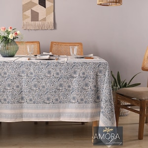 May include: A blue and white floral patterned tablecloth with a white border. The tablecloth is laid on a wooden table with four chairs. There is a woven wall hanging above the table and a woven light fixture hanging from the ceiling.