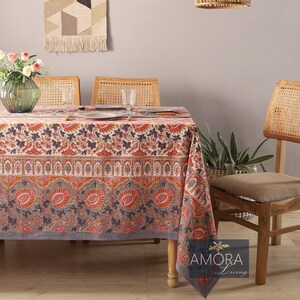 Puede incluir: Un mantel de mesa floral gris y rojo con un patrón paisley. El mantel está puesto sobre una mesa de madera con cuatro sillas. Hay un tapiz de pared tejido y una lámpara colgante de mimbre en el fondo.