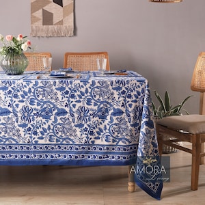 Puede incluir: Un mantel de mesa floral azul y blanco con un borde azul. El mantel está puesto sobre una mesa de madera con cuatro sillas. Hay un tapiz tejido en la pared sobre la mesa y una lámpara colgante de mimbre colgando del techo.