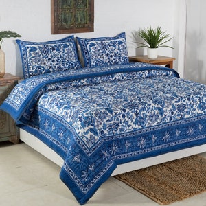 May include: A blue and white floral patterned quilt set on a bed. The set includes a quilt and two pillow shams. The quilt and shams feature a detailed floral design with a blue border. The bed is made up with a white sheet.