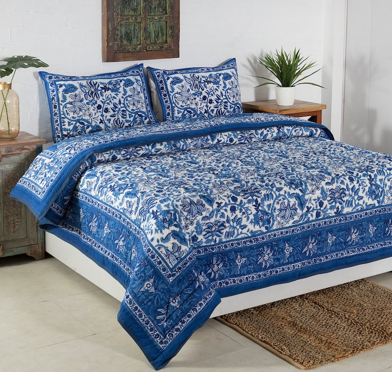 May include: A blue and white floral patterned quilt set on a bed. The set includes a quilt and two pillow shams. The quilt and shams feature a detailed floral design with a blue border. The bed is made up with a white sheet.