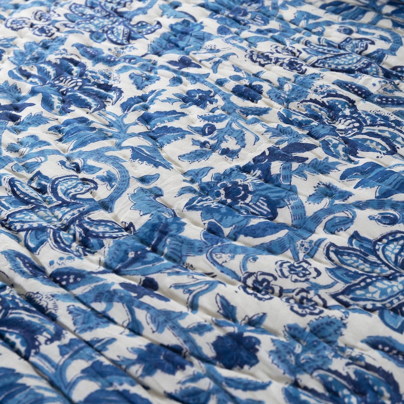 May include: A quilted textile featuring a repeating floral pattern in shades of blue on a white background. The design includes stylized flowers and foliage, with a textured, stitched appearance. The overall aesthetic is reminiscent of traditional block printing.