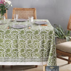Pode incluir: Uma toalha de mesa branca com um padrão floral verde e uma borda verde. A toalha de mesa está posta sobre uma mesa de madeira com duas cadeiras. Há pratos e copos na mesa.