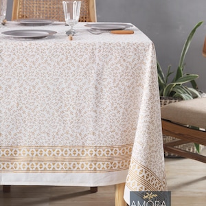 May include: White tablecloth with a gold leaf pattern and a gold geometric border. The tablecloth is laid on a wooden table with two place settings and a glass of water.
