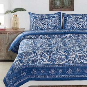 May include: A blue and white floral patterned quilt set on a bed. The set includes a quilt and two pillow shams. The quilt and shams feature a detailed floral design in shades of blue on a white background. The bed is made up with a woven rug.