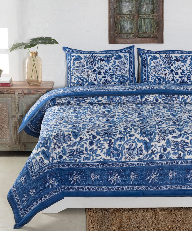 May include: A blue and white floral patterned quilt set on a bed. The set includes a quilt and two pillow shams. The quilt and shams feature a detailed floral design in shades of blue on a white background. The bed is made up with a woven rug.