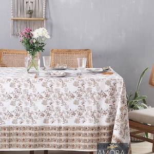 May include: White tablecloth with a brown floral pattern and a brown border. The tablecloth is laid on a wooden table with two chairs. There are flowers in a vase on the table. The text "AMORA Living" is visible in the bottom right corner of the image.