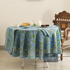 May include: A blue and green floral patterned tablecloth with a white border. The tablecloth is laid on a round table with a white base. There are two cups, a teapot, and a plate of cookies on the table. The text "AMORA Living" is visible in the lower right corner of the image.
