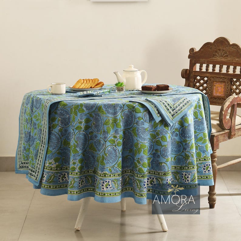 May include: A blue and green floral patterned tablecloth with a white border. The tablecloth is laid on a round table with a white base. There are two cups, a teapot, and a plate of cookies on the table. The text "AMORA Living" is visible in the lower right corner of the image.