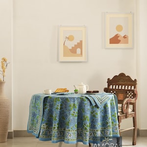 May include: A blue and green floral patterned tablecloth with a white border, covering a round table with a white base. The table is set with a teapot, cups, and plates. A wooden chair with intricate carvings is in the background.