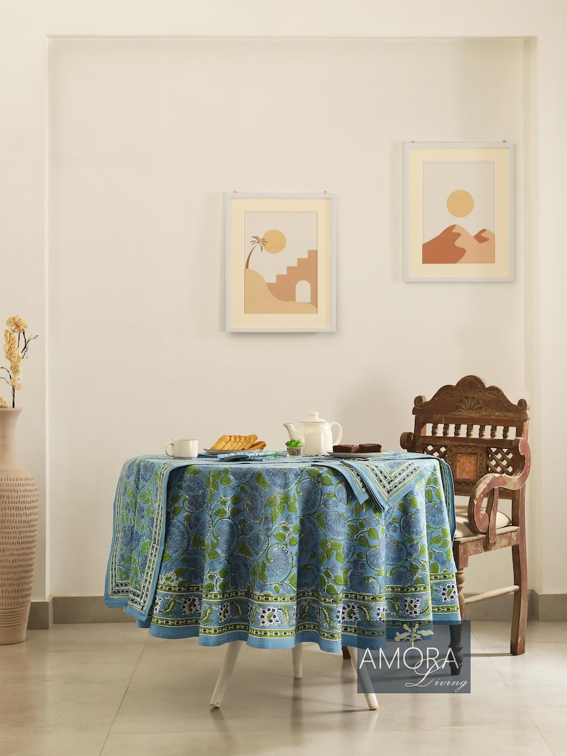 May include: A blue and green floral patterned tablecloth with a white border, covering a round table with a white base. The table is set with a teapot, cups, and plates. A wooden chair with intricate carvings is in the background.