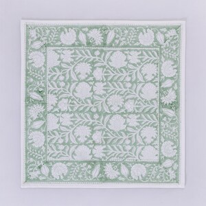 Pode incluir: Um tapete de mesa quadrado de tecido verde e branco com um padr&atilde;o floral. O tapete de mesa tem uma borda branca com um design floral.