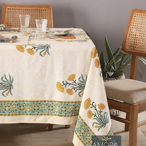Könnte beinhalten: Eine weiße Tischdecke mit einem floralen Muster in Gelb-, Grün- und Blautönen. Die Tischdecke hat einen Rand mit einem sich wiederholenden Muster aus kleinen Blumen. Die Tischdecke liegt auf einem Holztisch mit zwei Stühlen.