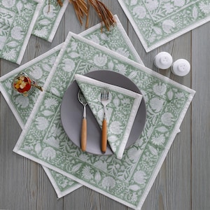 Pode incluir: Um conjunto de quatro  placemats verdes e brancos com padr&atilde;o floral, com um prato cinza, dois garfos de prata e uma colher de prata sobre uma toalha de mesa branca. Os placemats s&atilde;o feitos de tecido e t&ecirc;m uma borda branca.