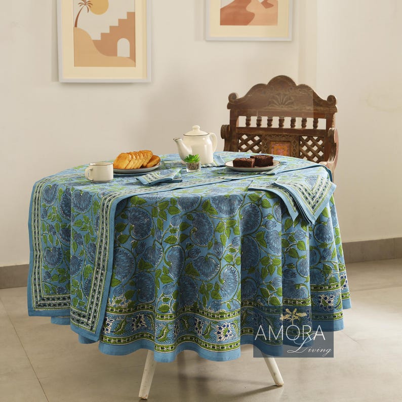 May include: A blue and green floral round tablecloth with a white border. The tablecloth is set on a white table with a wooden chair and a teapot, snacks, and a plate of brownies on the table. The text "AMORA Living" is visible in the bottom right corner of the image.