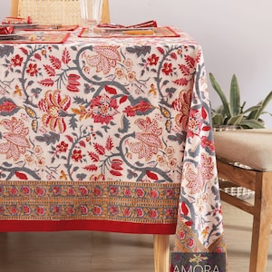 Puede incluir: Mantel floral crema con borde rojo. El diseño presenta flores rojas, rosas y amarillas con tallos y hojas grises. La mesa está puesta con servilletas y manteles individuales a juego, creando un ambiente acogedor.