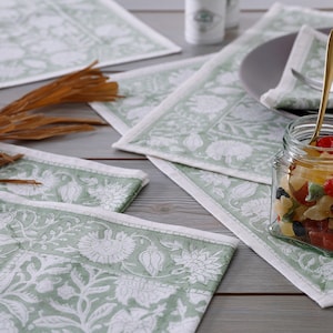 Pode incluir: Conjunto de quatro  placemats com padr&atilde;o floral verde e branco. Os placemats s&atilde;o feitos de algod&atilde;o e t&ecirc;m um brilho sutil. Os placemats est&atilde;o dispostos em uma mesa de madeira.