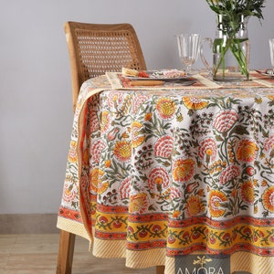以下が含まれることがあります： オレンジ、イエロー、グリーンの色合いの花柄が施された、丸い白いテーブルクロス。テーブルクロスは、ストライプ模様のイエローとオレンジの縁取りが施されています。テーブルクロスは、椅子と花瓶のある木製のテーブルの上に置かれています。
