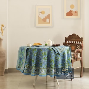 May include: A blue and green floral patterned tablecloth with a white border, set on a round table with a white base. The table is set with two cups, a plate of cookies, and a teapot. A wooden chair with a carved back is in the background.