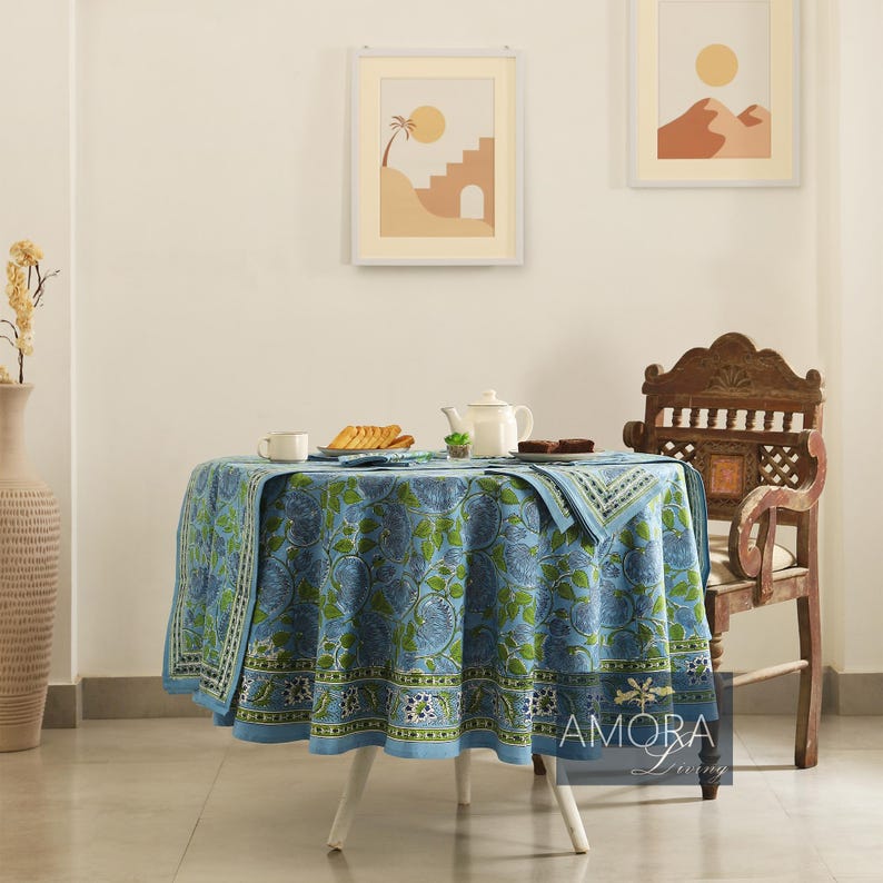 May include: A blue and green floral patterned tablecloth with a white border, set on a round table with a white base. The table is set with two cups, a plate of cookies, and a teapot. A wooden chair with a carved back is in the background.