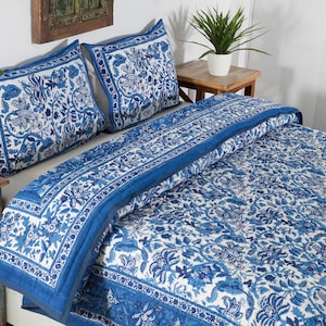 May include: A blue and white floral patterned quilt and two matching pillow shams rest on a bed. The bedding features a detailed floral design with a solid blue border. The room has a rustic, bohemian aesthetic.