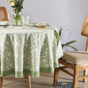 Puede incluir: Un mantel de mesa redondo verde y blanco con flores y un borde verde, colocado sobre una mesa redonda de madera con dos sillas de madera. La mesa está puesta con un jarrón de flores, vasos y un plato.
